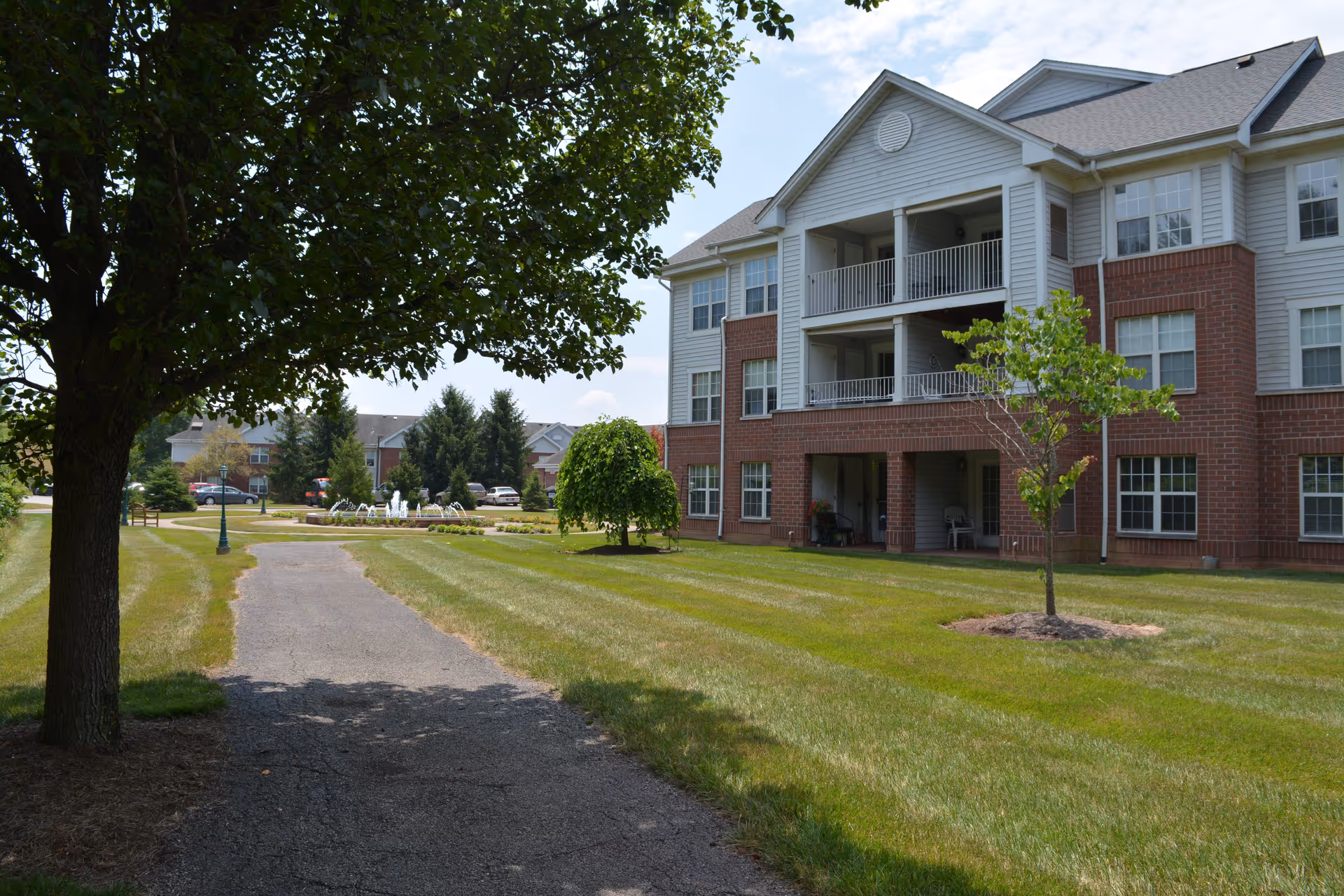 Exterior view of a multi-story residential building with balconies and a brick and siding facade, surrounded by a well-maintained lawn, trees, and a paved walkway leading towards a fountain in the distance under a partly cloudy sky.
