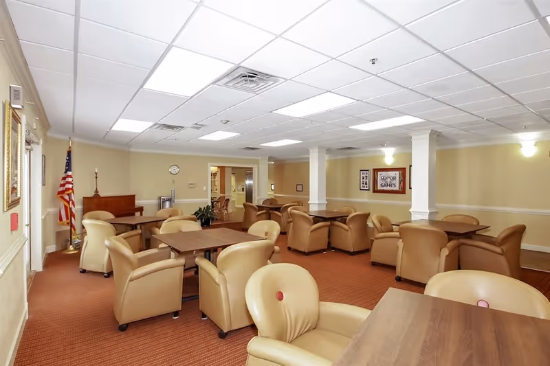 A spacious room with multiple wooden tables surrounded by beige cushioned chairs on a red carpeted floor. The walls are light yellow with white trim, and there are white columns dividing the space. An American flag stands in the corner near a piano. The ceiling has white tiles with recessed lighting. A framed sign on the wall reads 'Senior Games'.