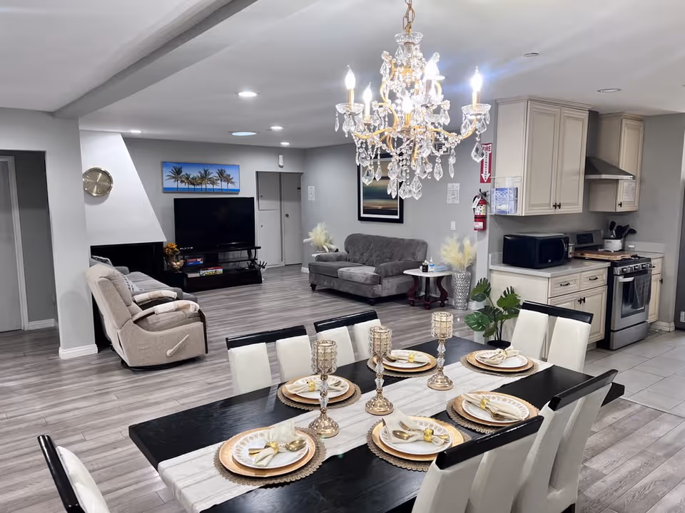 Open-plan interior showing a set dining table under a crystal chandelier with a living area and kitchen in the background.