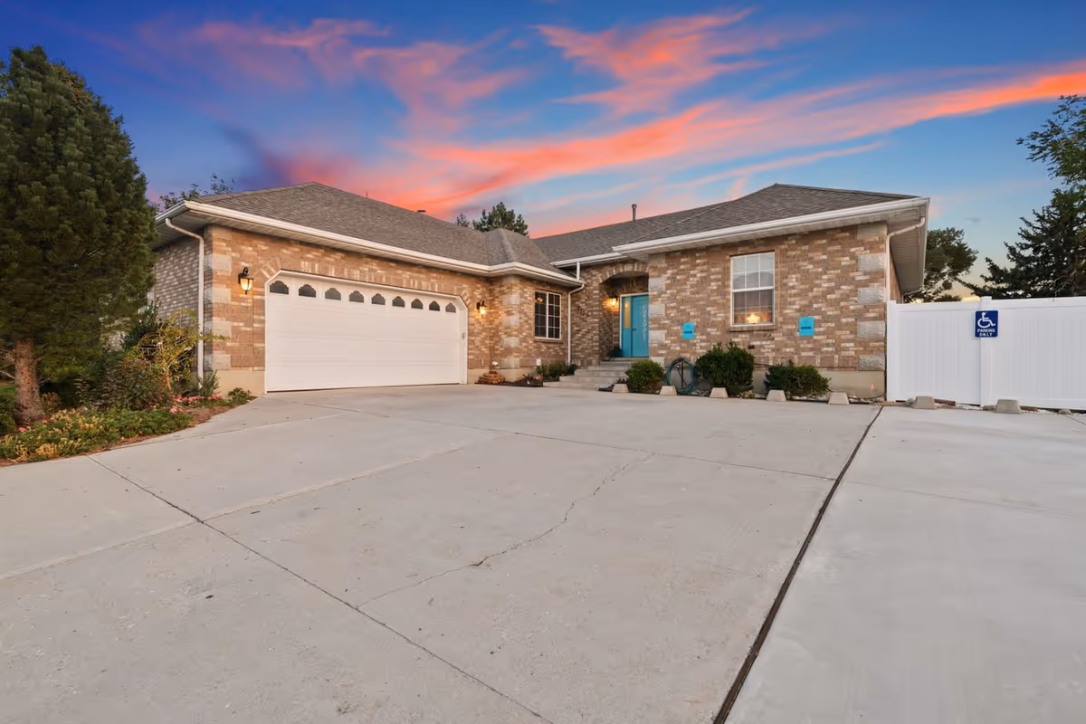 Exterior view of a single-story brick residential building with a two-car garage, a turquoise front door, and a concrete driveway. There are small bushes near the entrance and a white fence with a handicap parking sign. The sky is colorful with a sunset glow.