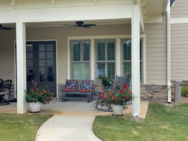 Covered outdoor patio area with a concrete floor, featuring a bench with red cushions and patterned pillows, two rocking chairs with red cushions, and two large potted plants with red flowers. The patio is attached to a beige building with white trim and has a ceiling fan. A curved concrete walkway leads up to the patio through a grassy lawn.