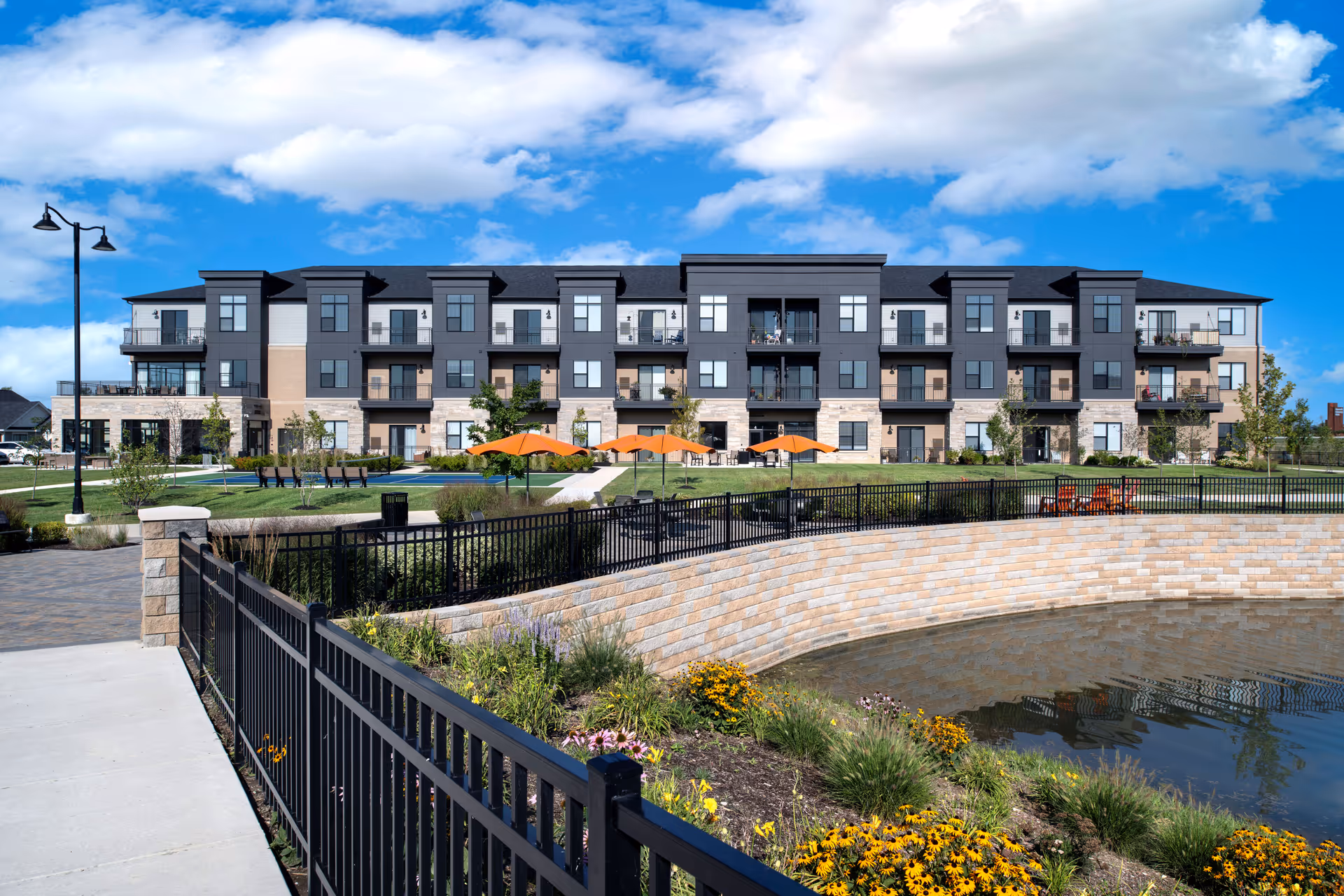A modern three-story senior living facility building with balconies, surrounded by landscaped gardens, orange patio umbrellas, benches, and a small pond with a stone retaining wall under a partly cloudy blue sky.