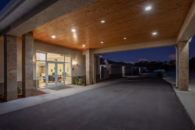 Entrance area of a senior living facility at night with a covered driveway, illuminated ceiling lights, glass double doors leading inside, and an American flag visible in the background.
