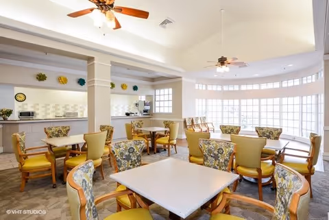 Bright dining room with multiple square tables surrounded by chairs with floral and yellow upholstery. The room features large windows allowing natural light, ceiling fans with lights, and a counter area with a coffee machine and microwave in the background.