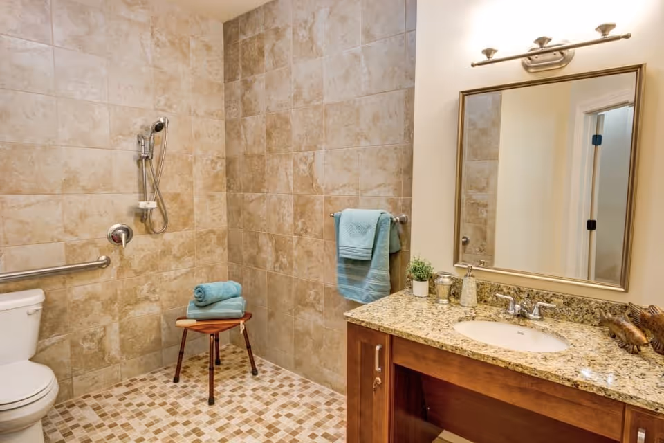 A bathroom with beige tiled walls and floor, featuring a walk-in shower with a handheld showerhead, a white toilet with a grab bar, a small wooden stool with folded blue towels, a granite countertop with a sink, a large mirror above the sink, and a towel rack holding blue towels.