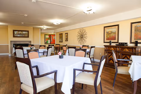 Bright dining room with tables covered in white tablecloths, chairs, wall art and wood flooring.