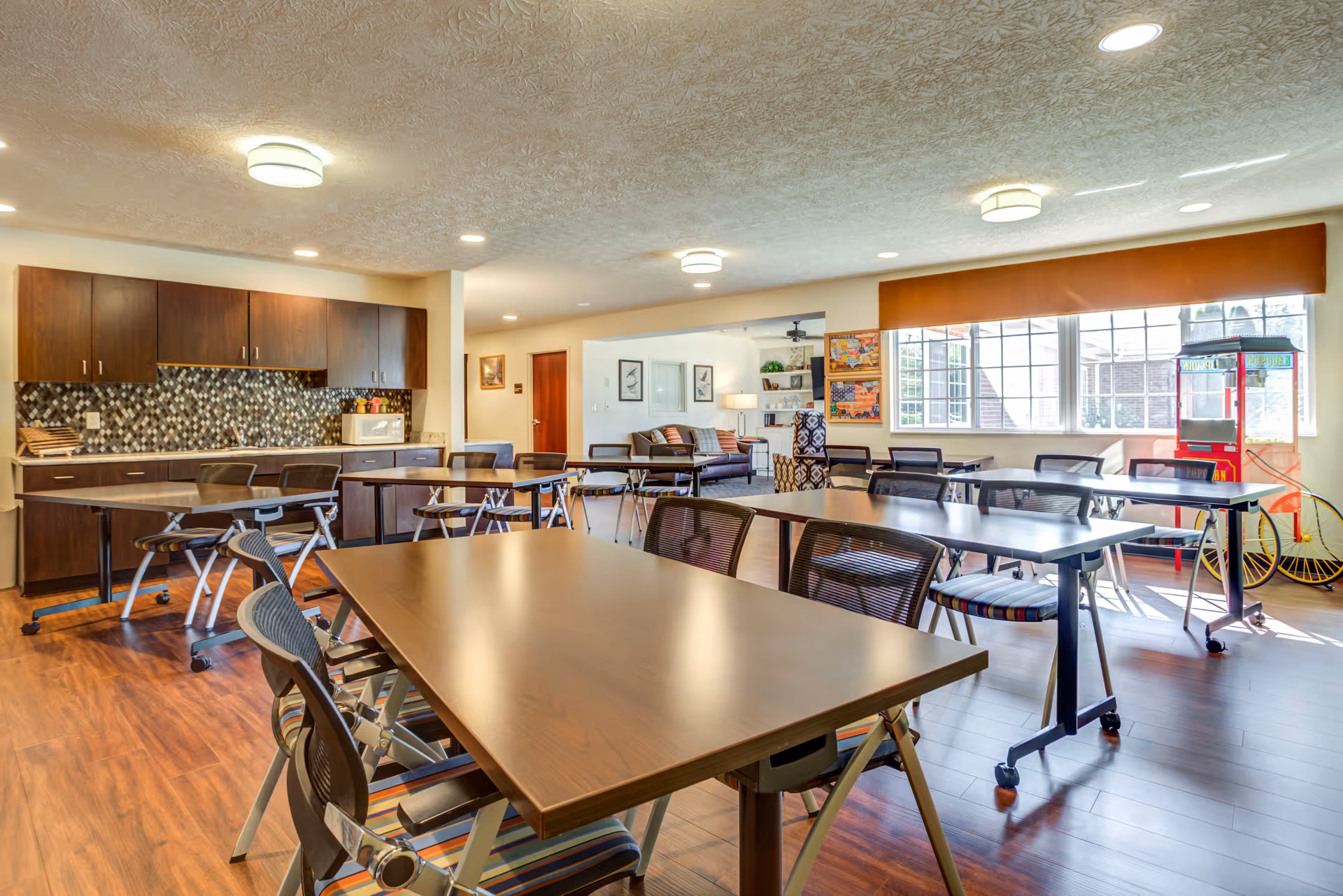 A bright and spacious common area in Glen Ridge Health Campus featuring several tables and chairs arranged for group activities or dining. The room has large windows letting in natural light, a kitchenette with dark wood cabinets and a mosaic tile backsplash, and a cozy seating area with couches and framed artwork on the walls. There is also a popcorn machine near the windows.