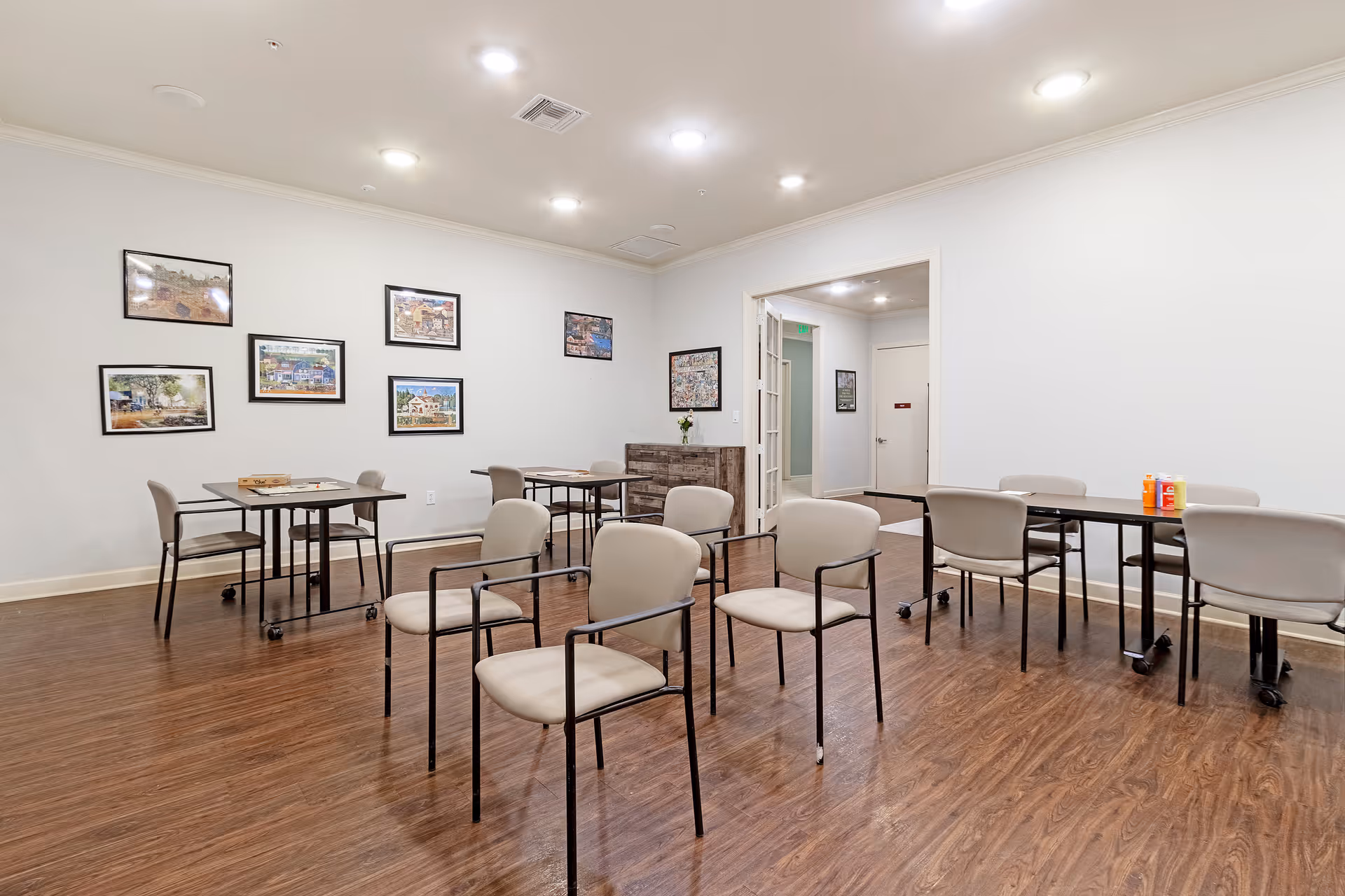 A well-lit room with wooden flooring featuring several beige chairs with black metal frames arranged around black tables on wheels. The walls are white and decorated with framed pictures. There is an open doorway leading to a hallway with additional framed pictures on the walls.
