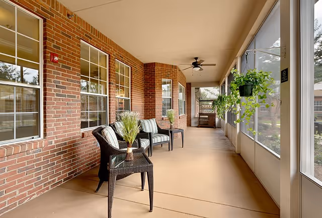 A screened-in porch area with brick walls and multiple windows on one side, and large screened panels on the other. The porch is furnished with black wicker chairs with striped cushions and small tables holding potted plants. Ceiling fans are mounted on the white ceiling, and hanging plants are suspended from the screened panels.