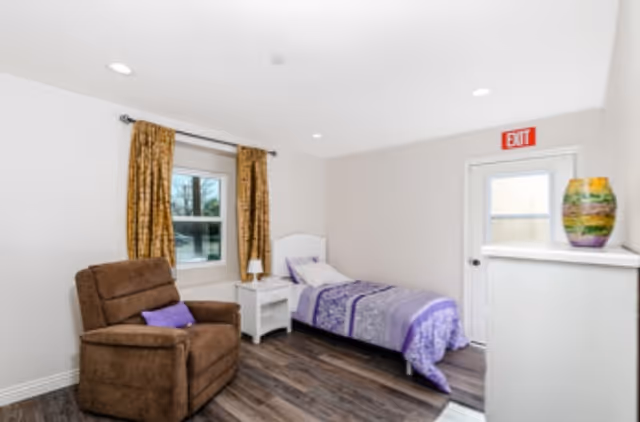 A small, cozy bedroom with a single bed covered in purple bedding, a brown armchair with a purple pillow, a white nightstand with a lamp, a window with gold curtains, and a white dresser with a colorful vase on top. There is an exit door with a red exit sign above it.