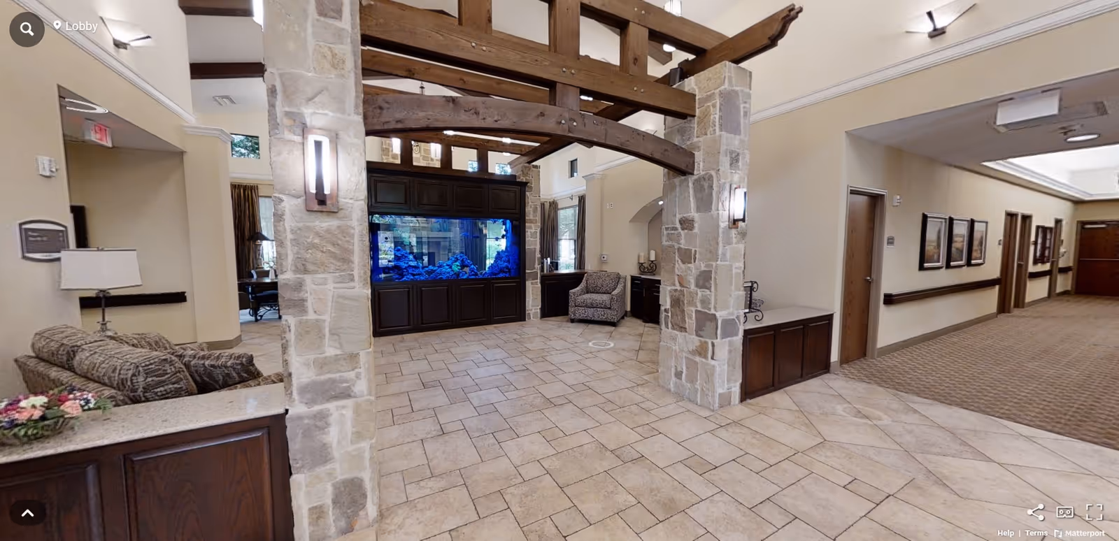 Lobby area with stone pillars and wooden beams overhead, featuring a large built-in aquarium with blue lighting. There is a patterned sofa on the left, an armchair near the aquarium, and a hallway with framed pictures on the right. The floor is tiled and the walls are painted beige.