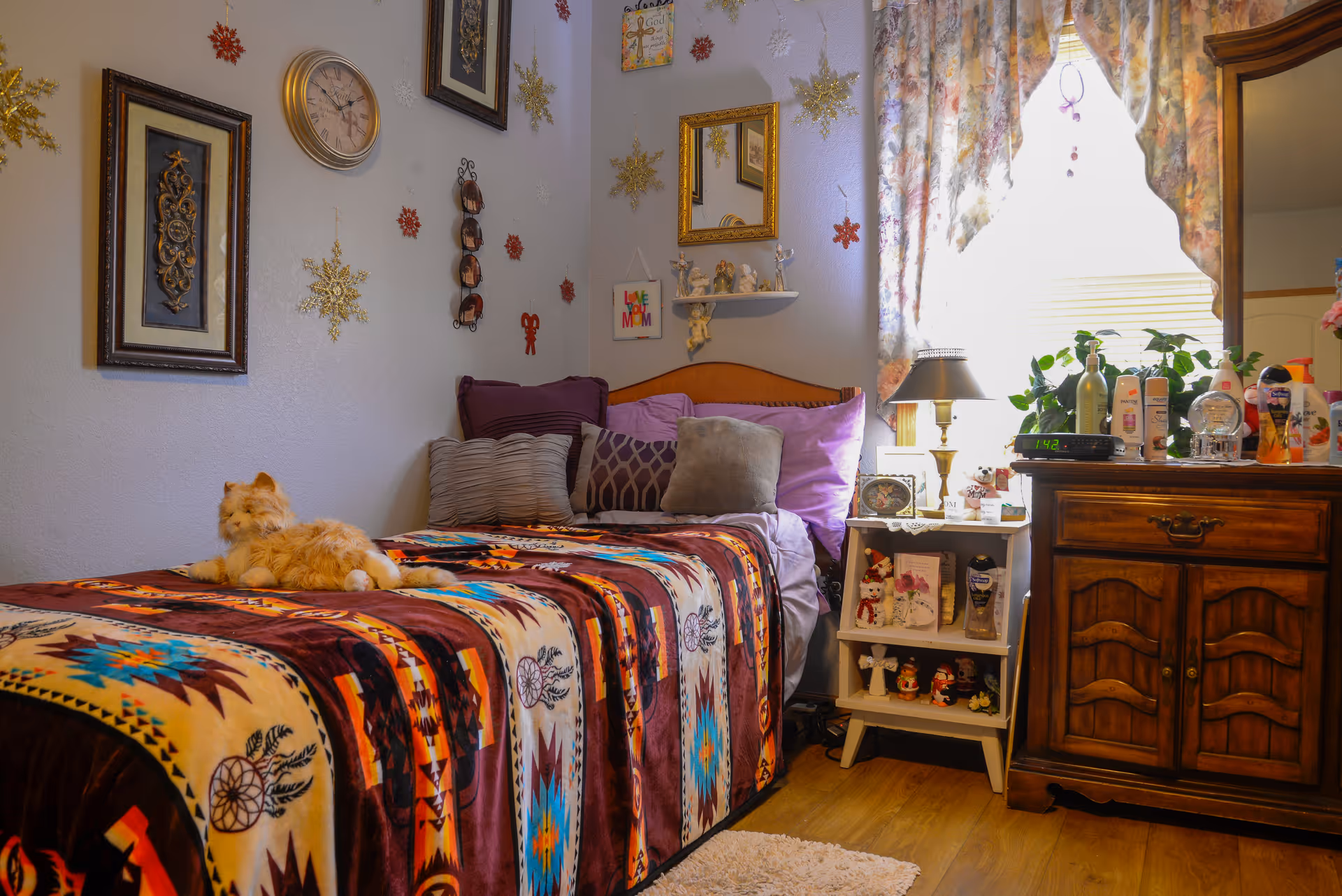 Cozy decorated bedroom featuring a single bed with a colorful patterned blanket and stuffed cat, pillows, a nightstand with lamp and decorations, and a wooden dresser by a curtained window.