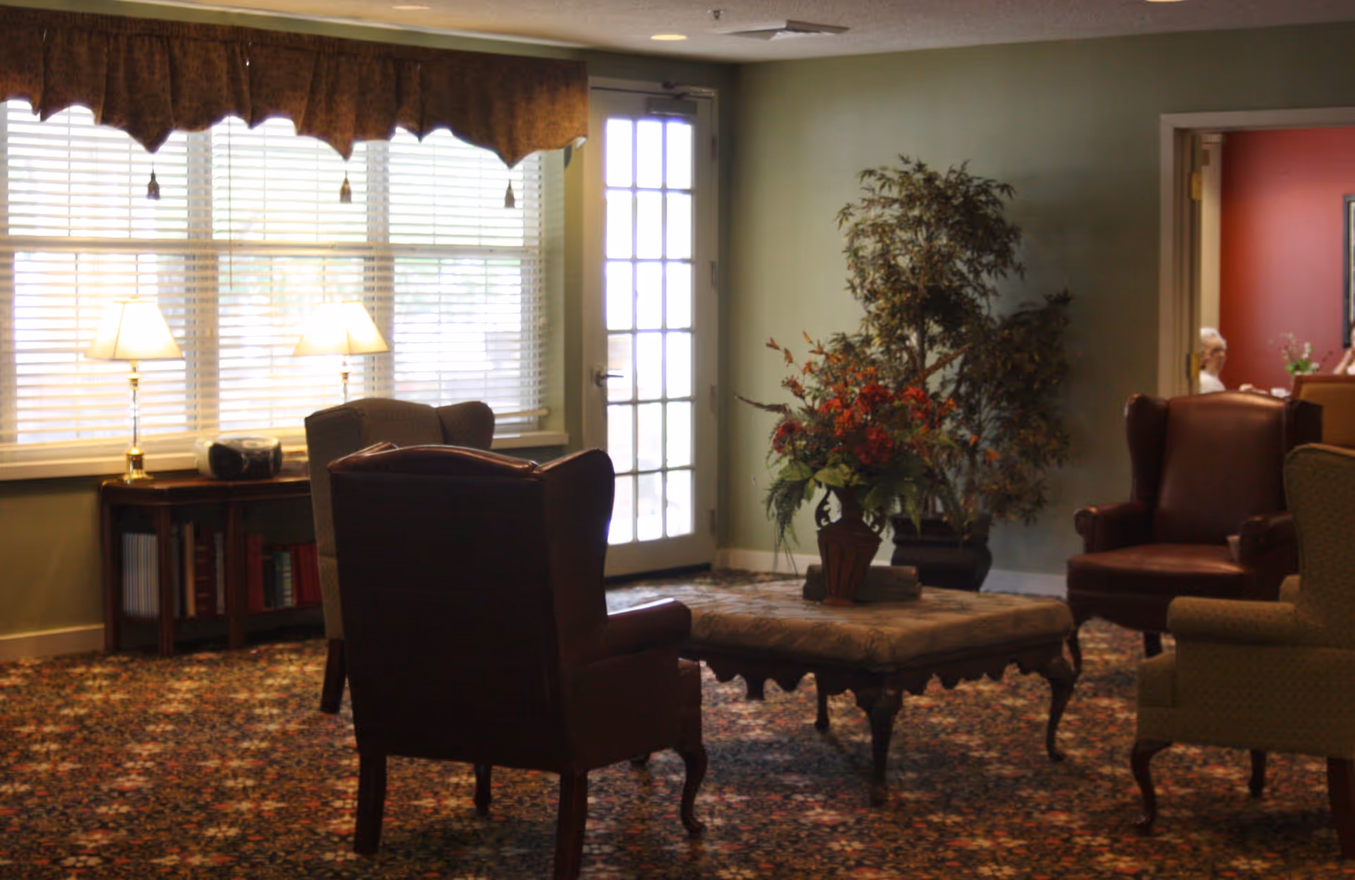 A cozy senior living common area with several upholstered armchairs arranged around a large ottoman topped with a floral arrangement. The room has a patterned carpet, a window with blinds and valance, two table lamps on a side table, and a glass door leading outside. There is a large potted plant in the corner and a glimpse of another room with red walls and a person sitting inside.