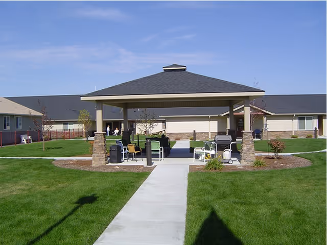 Outdoor covered seating area with chairs and tables on a concrete patio surrounded by green grass and pathways, with a single-story building in the background under a clear blue sky.