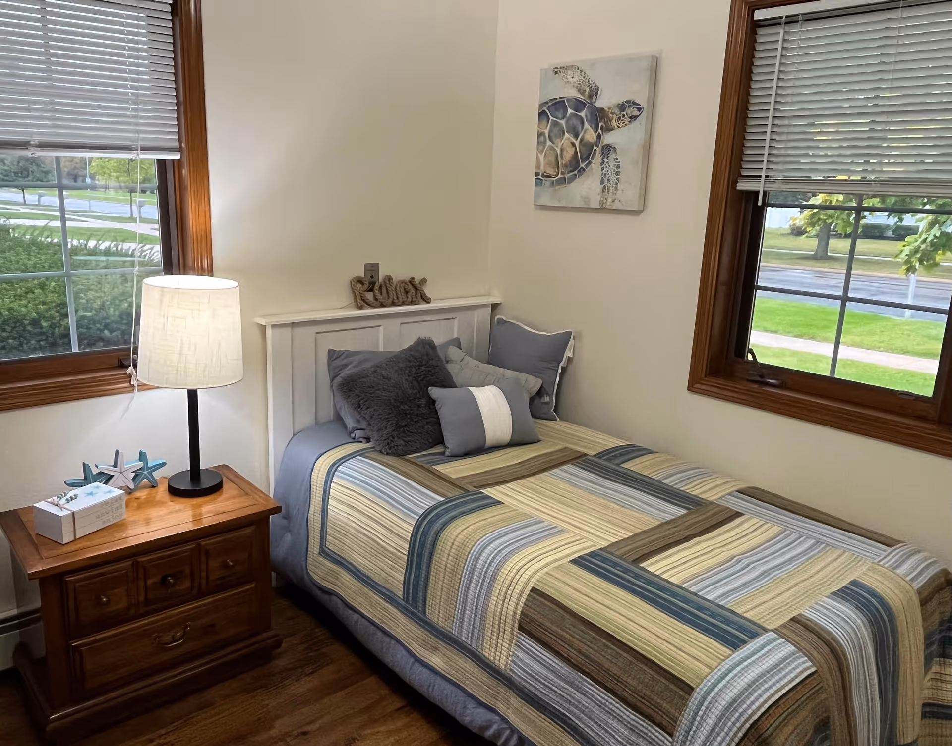A cozy bedroom with a single bed covered in a striped quilt in shades of blue, green, and beige. The bed has multiple pillows including a furry gray one and a cylindrical bolster pillow. There is a wooden nightstand next to the bed with a lamp, decorative starfish, and a small box. Two windows with wooden frames and blinds let in natural light. A painting of a sea turtle hangs on the wall above the bed.