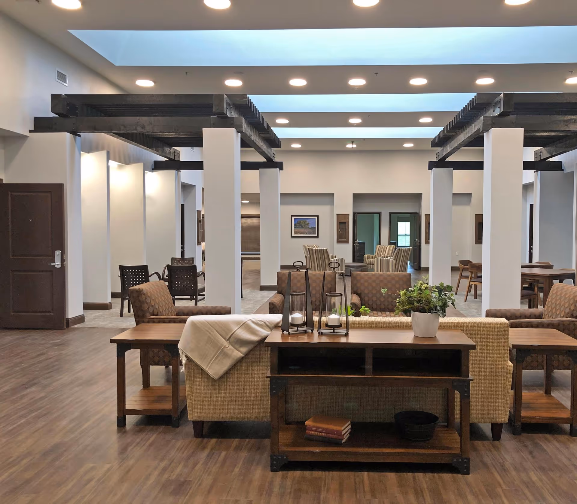 A spacious and well-lit common area in a retirement community featuring multiple seating arrangements with armchairs and sofas. The room has wooden flooring, white walls, and a ceiling with recessed lighting and skylights. There are decorative elements such as plants and candle holders on wooden tables, and framed artwork on the walls.