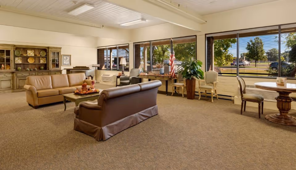 A spacious and well-lit living room area in a senior living facility with large windows showing an outdoor view. The room features two leather sofas facing each other with a coffee table in between, a wooden cabinet with decorative items, a piano, several chairs, a small round table with chairs, and an American flag near the windows.