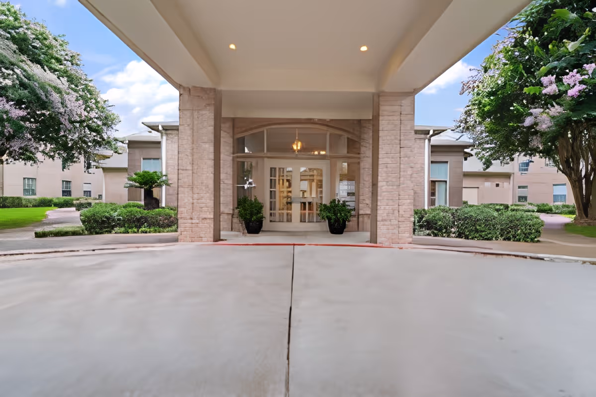 Covered porte-cochere entrance leading to glass double doors of a senior living facility with potted plants and landscaped grounds.