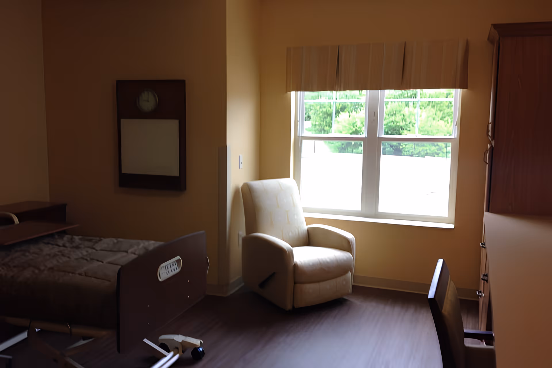 A senior living bedroom with a medical-style bed, a recliner by a bright window, and wooden furniture.