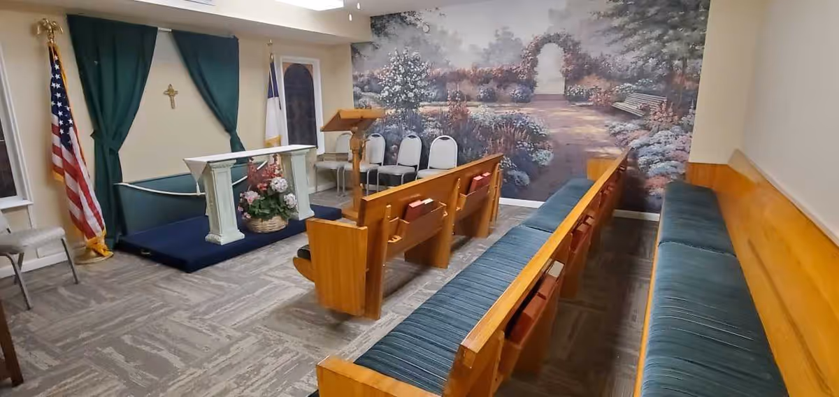Small chapel interior with wooden pews, an altar, flags, and a pastoral mural on the back wall.