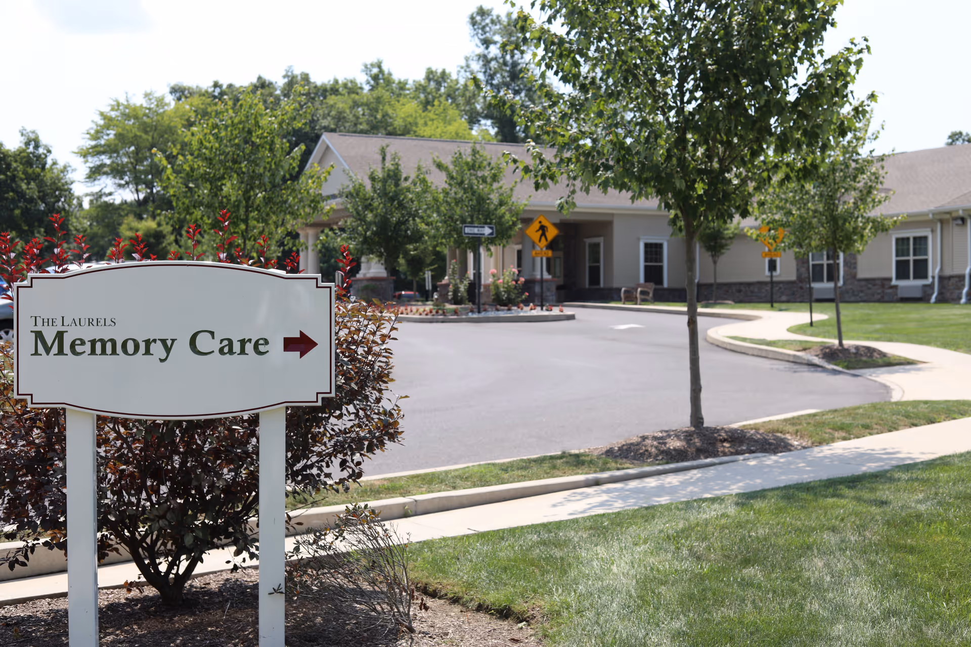 Outdoor view of a senior living facility with a sign in the foreground that reads 'The Laurels Memory Care' with an arrow pointing right. The background shows a paved driveway, trees, and a beige building with a covered entrance.