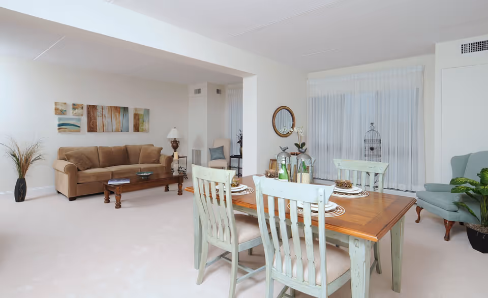 A bright and spacious living and dining area in a senior living facility. The room features a beige sofa with a wooden coffee table in front, a wooden dining table with four light green chairs, and a green armchair near a large window with sheer white curtains. Decorative items include wall art above the sofa, a round mirror on the wall, a birdcage, a table lamp, and potted plants.