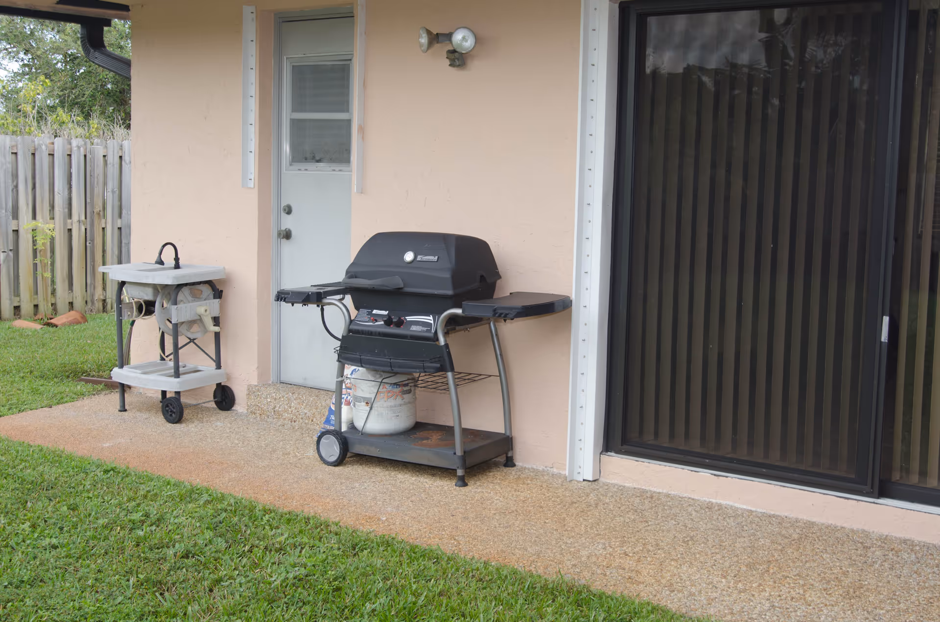 Outdoor patio area with a gas grill and a small utility cart next to a beige exterior wall with a white door and a sliding glass door with vertical blinds. There is a grassy lawn and a wooden fence in the background.