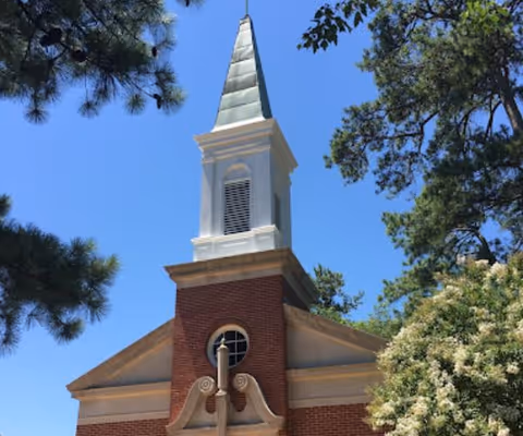 The steeple of a brick building with a white and green pointed top, surrounded by green trees under a clear blue sky.