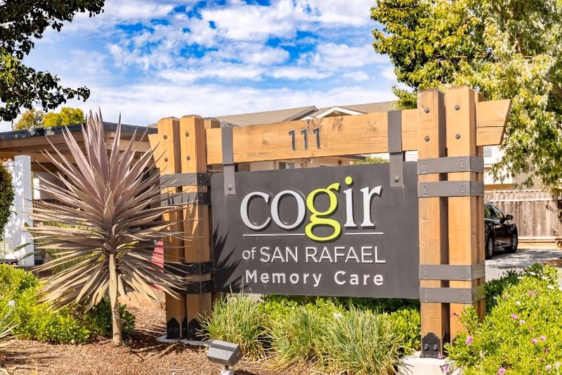 Outdoor wooden sign for Cogir of San Rafael Memory Care with the address number 111 above it, surrounded by plants and trees under a partly cloudy sky.