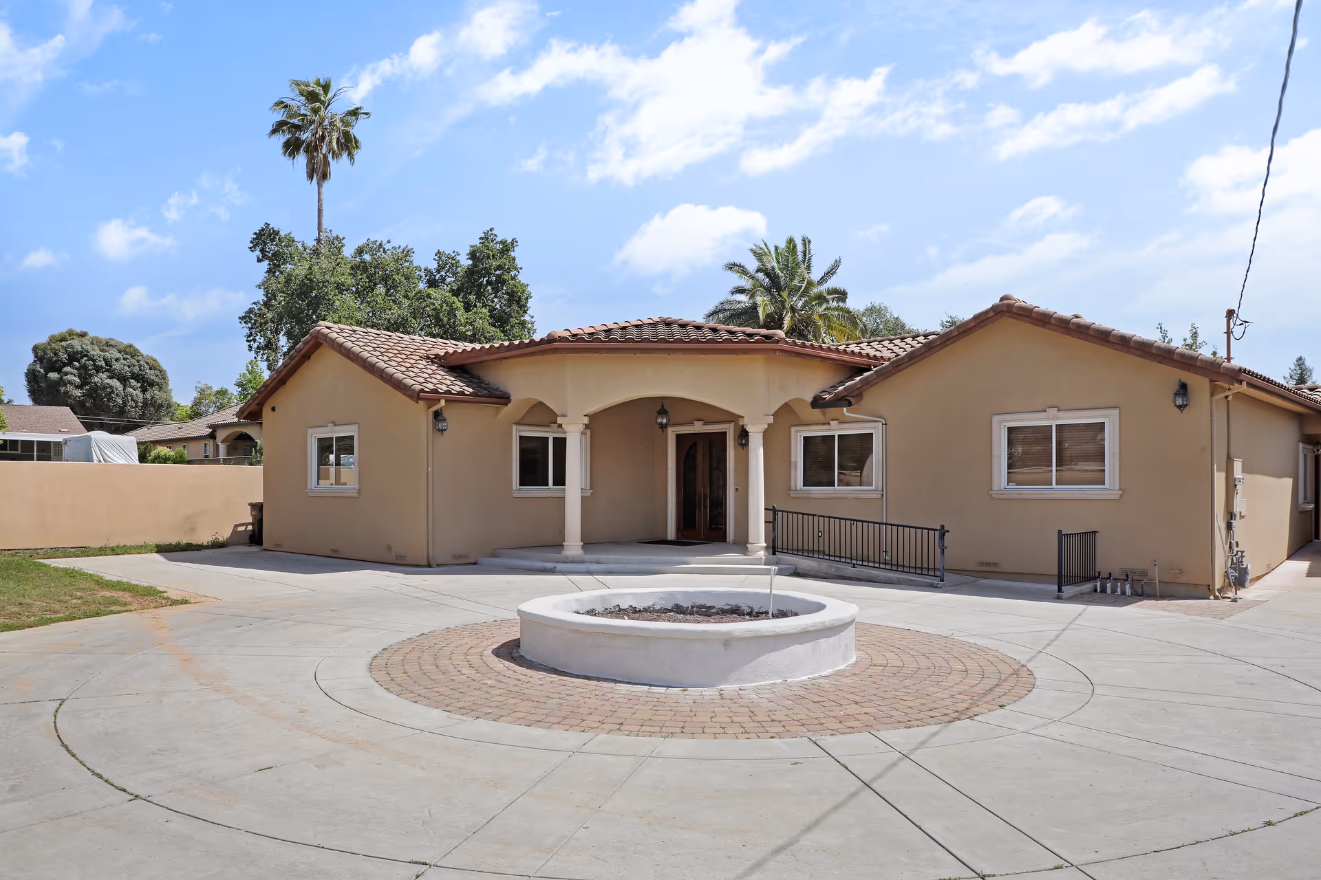 Front exterior view of a single-story beige building with a tiled roof, an arched entrance supported by two columns, and a circular driveway with a raised round planter in the center. There are palm trees and other greenery in the background under a partly cloudy blue sky.