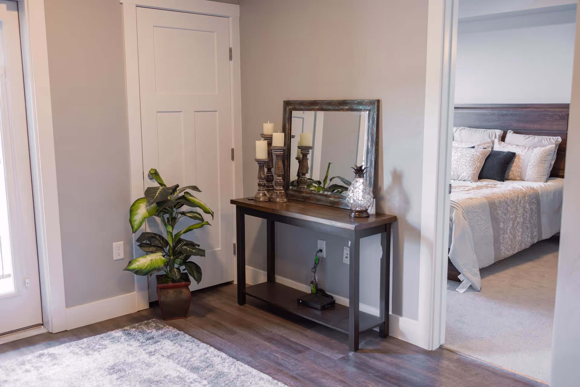Interior view of a hallway corner featuring a wooden console table with a mirror, decorative candles, and a glass pineapple ornament. A potted plant is placed beside a closed white door. To the right, an open doorway reveals a bedroom with a bed made up with patterned bedding and multiple pillows.