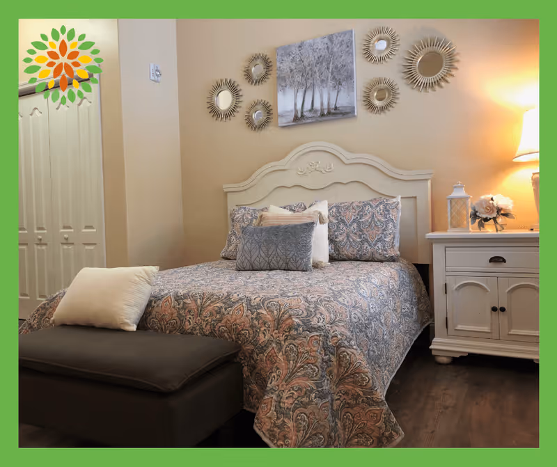 A cozy bedroom with a patterned bedspread on a bed with a decorative white headboard. There are multiple pillows on the bed, a dark cushioned bench at the foot of the bed, and a white nightstand with a lamp, a lantern, and a flower arrangement. The wall above the bed features a painting of trees and several round decorative mirrors.