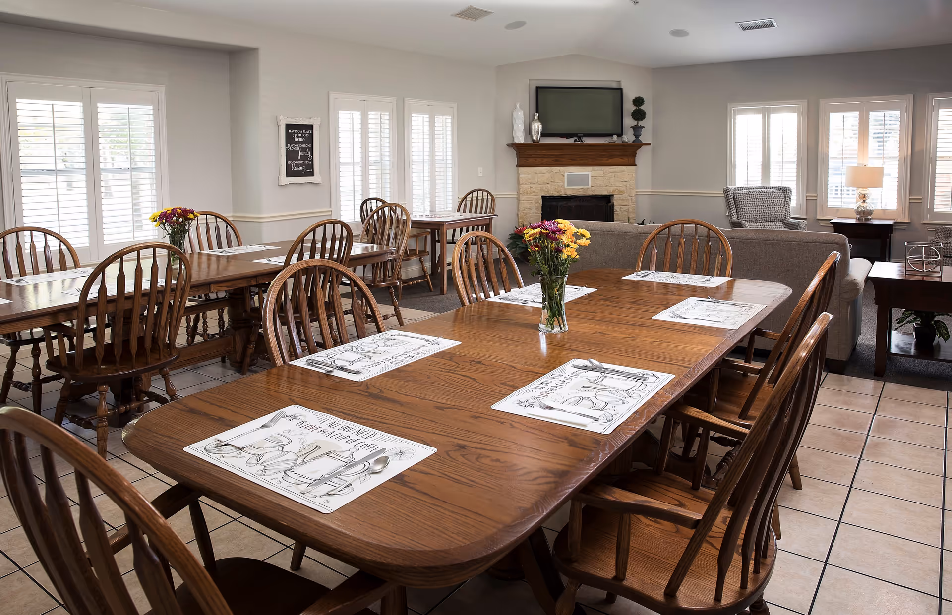 A bright dining room with wooden tables and chairs arranged neatly. Each table has placemats and cutlery set, with a vase of colorful flowers as a centerpiece. The room features large windows with white shutters, a stone fireplace with a TV mounted above it, and a cozy seating area with a sofa and armchair near the windows.