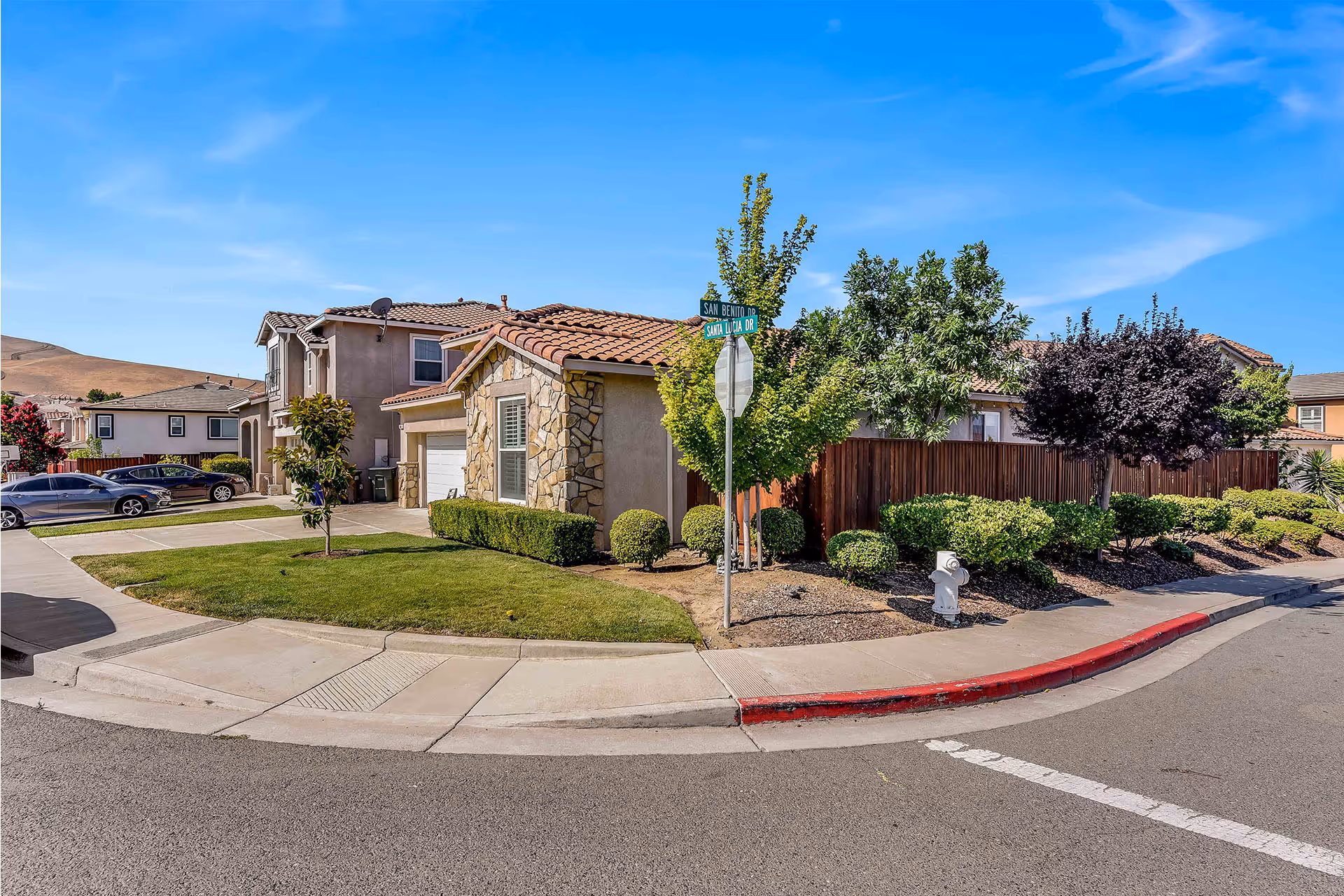 Street corner view of a residential neighborhood with houses, manicured lawns, trees, and parked cars under a clear blue sky.
