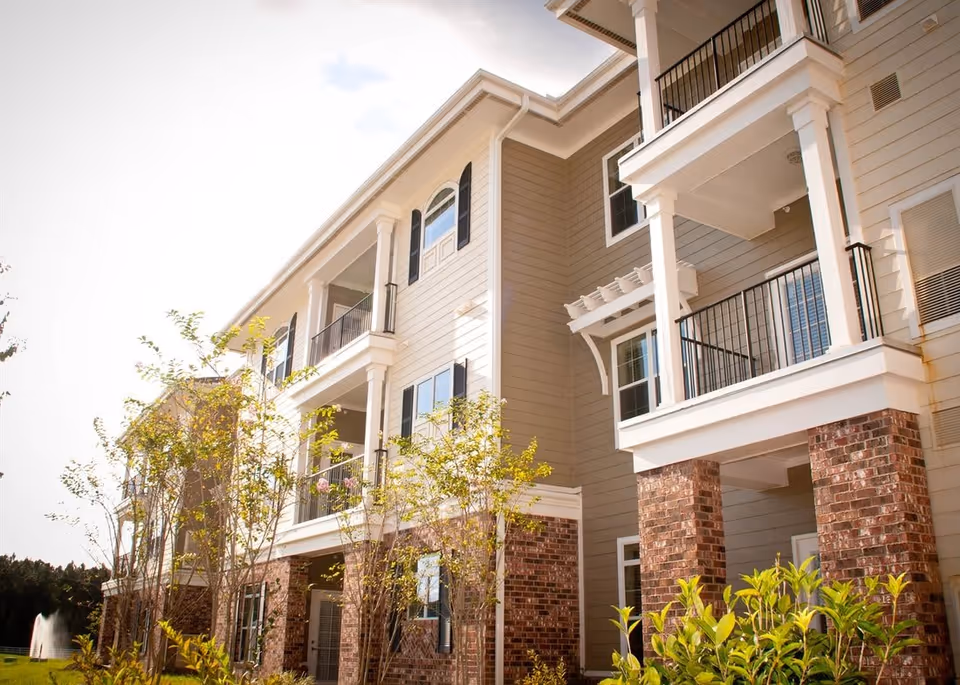 Exterior view of a multi-story retirement community building with beige siding, brick pillars, balconies with black railings, and small trees and shrubs in the foreground under a bright sky.