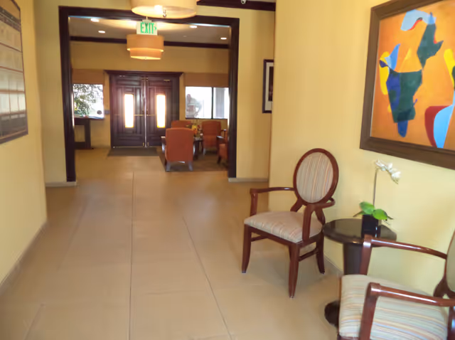 Interior hallway of an assisted living facility with beige tiled floor and yellow walls. Two wooden chairs with striped cushions are placed on the right side next to a small round table with a potted plant. A colorful abstract painting hangs above the chairs. At the end of the hallway, there is a seating area with orange armchairs and a table, and double wooden doors with glass panels. Ceiling lights and an exit sign are visible.