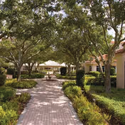 Brick-paved walkway through a tree-lined courtyard with manicured shrubs and a gazebo or fountain visible ahead.