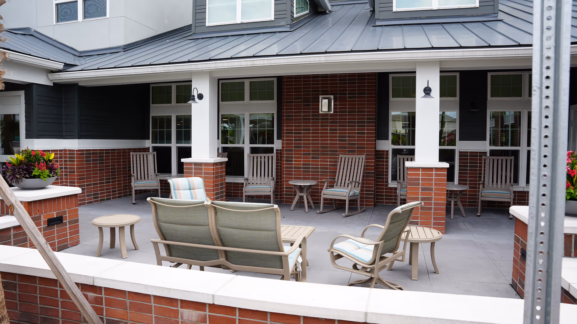Outdoor patio area at The Savoy Gracious Retirement Living featuring multiple cushioned chairs and small tables arranged on a concrete floor, surrounded by brick walls and white pillars with a metal roof overhead.