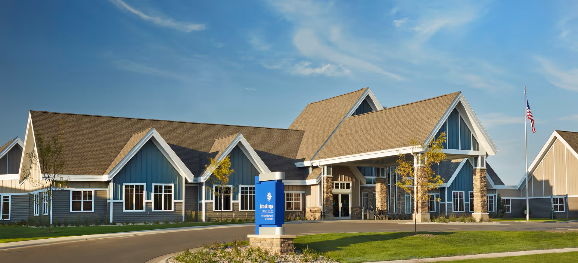 Exterior view of a large senior living facility building with blue and beige siding, multiple peaked roofs, and a covered entrance. There is a sign in front that reads 'Brookings' and an American flag on a flagpole to the right. The sky is clear with a few clouds.