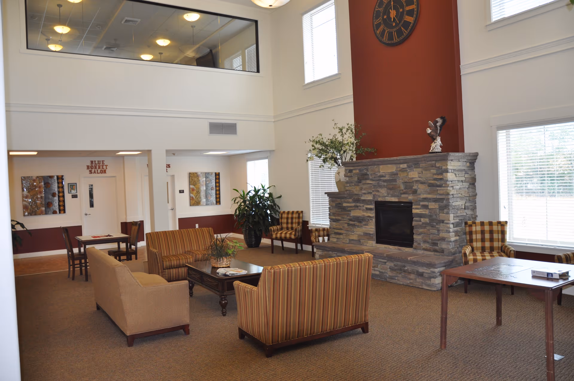 A spacious assisted living facility common area with a stone fireplace, a large clock above it, and several chairs and sofas arranged around a coffee table. There are large windows with blinds letting in natural light, a potted plant, and a small table with chairs in the background near a door labeled 'Blue Bonnet Salon'.
