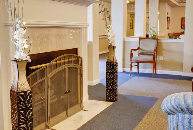 Interior view of a senior living facility lounge area featuring a fireplace with a decorative metal screen, two tall vases with white flowers on either side of the fireplace, a wooden chair with a cushion, and part of a blue patterned sofa. The space has beige walls and a carpeted floor with a mix of blue and beige tones.