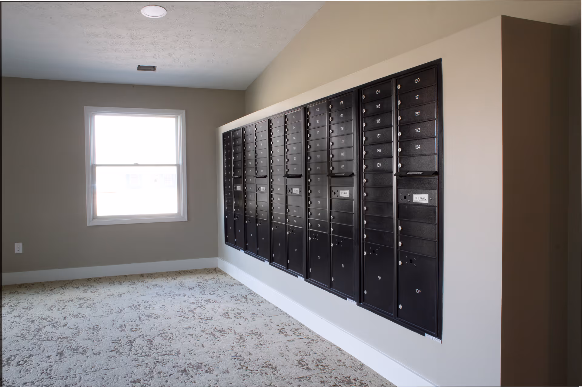 Interior room with a wall-mounted bank of black mailboxes numbered and labeled U.S. Mail, next to a window with natural light coming through. The room has beige walls and patterned carpet flooring.