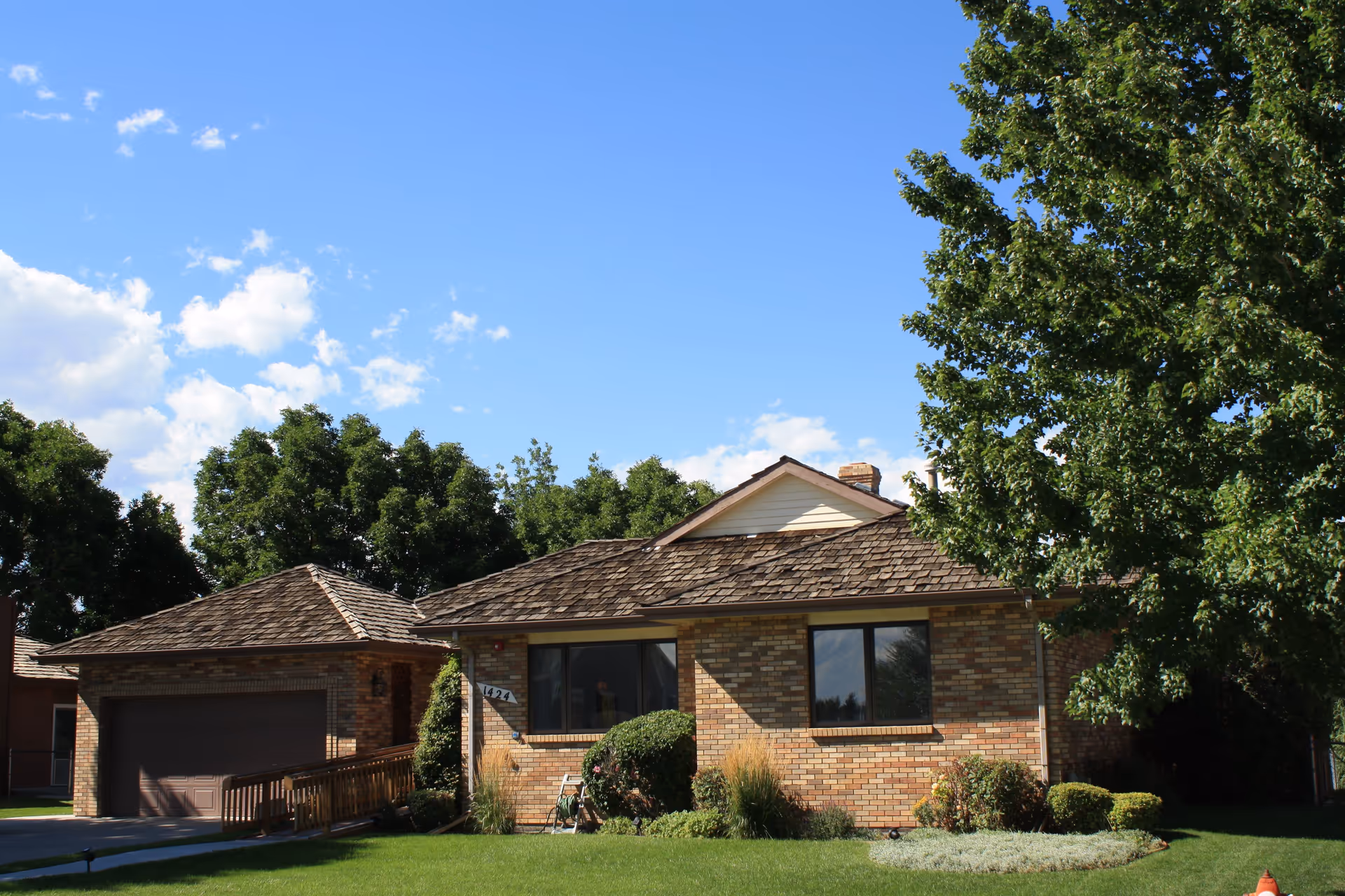 Single-story brick building with a shingled roof, two large windows, a garage with a brown door, a wooden ramp, and well-maintained green lawn and bushes under a clear blue sky with some clouds.