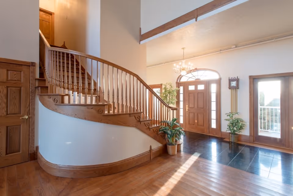 Bright entrance area of a senior living facility with a wooden curved staircase, wooden front door with glass panels, a chandelier, potted plants, and a wall clock.