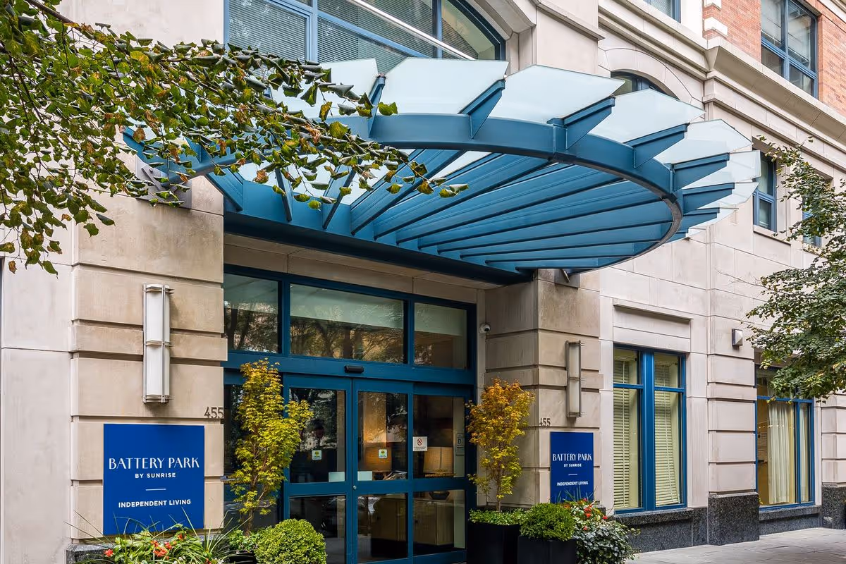 Entrance of Battery Park by Sunrise independent living facility with a blue glass canopy above the doorway, beige stone exterior walls, blue-framed windows, and green plants on either side of the entrance.