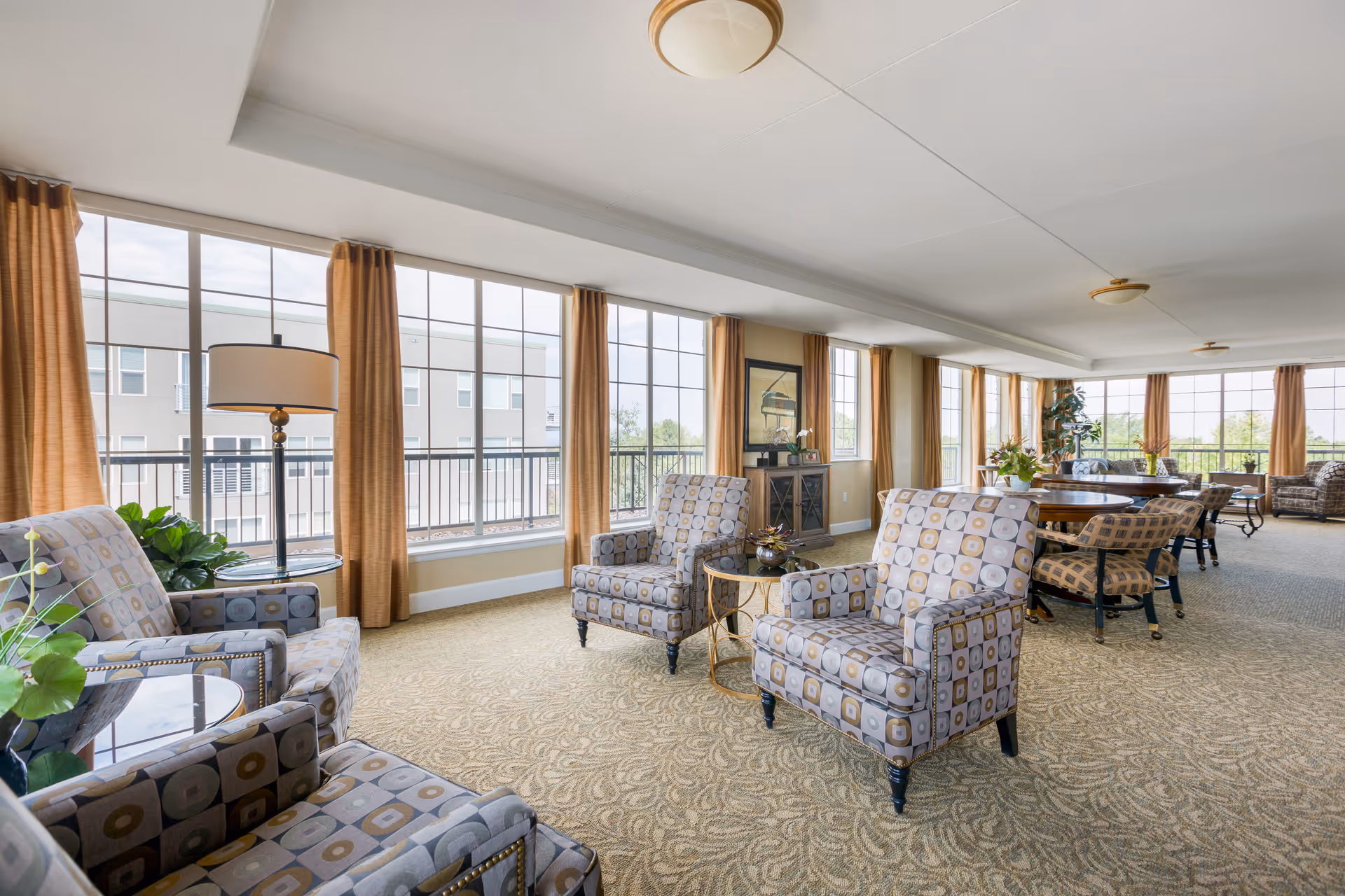 A spacious and well-lit living room area with large windows covered by orange curtains, patterned armchairs arranged around small tables, a round dining table with chairs, and decorative plants. The room has a beige patterned carpet and ceiling lights.
