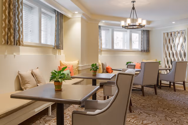A cozy interior common area with several tables and cushioned chairs arranged for seating. The room features beige walls, patterned carpet, windows with blinds and curtains, decorative pillows on bench seating, potted plants on tables, and a chandelier hanging from the ceiling.