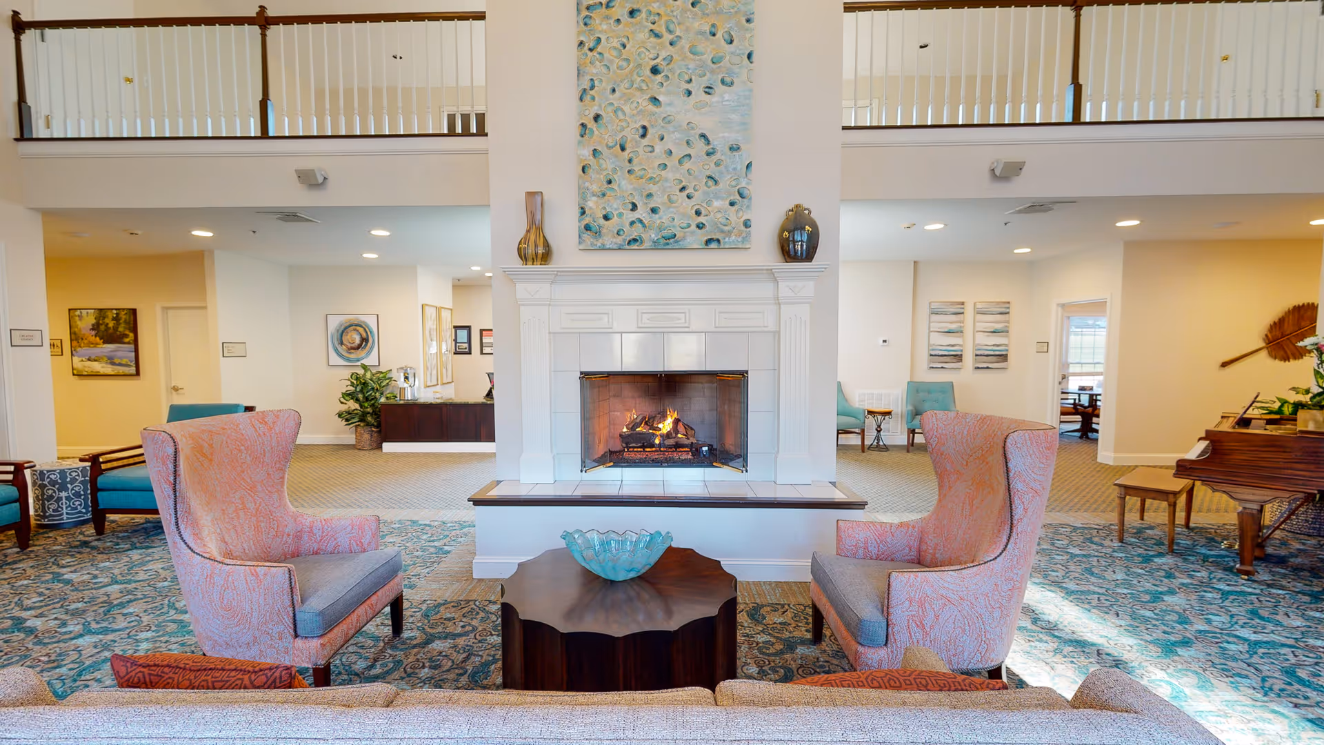 A spacious and well-lit living room area with a central white fireplace featuring a burning fire. Two patterned armchairs face the fireplace with a dark wooden coffee table between them, holding a decorative glass bowl. The room has a blue and beige patterned carpet, light-colored walls, and various pieces of artwork on the walls. A grand piano is visible on the right side, and a balcony railing runs along the upper part of the room.
