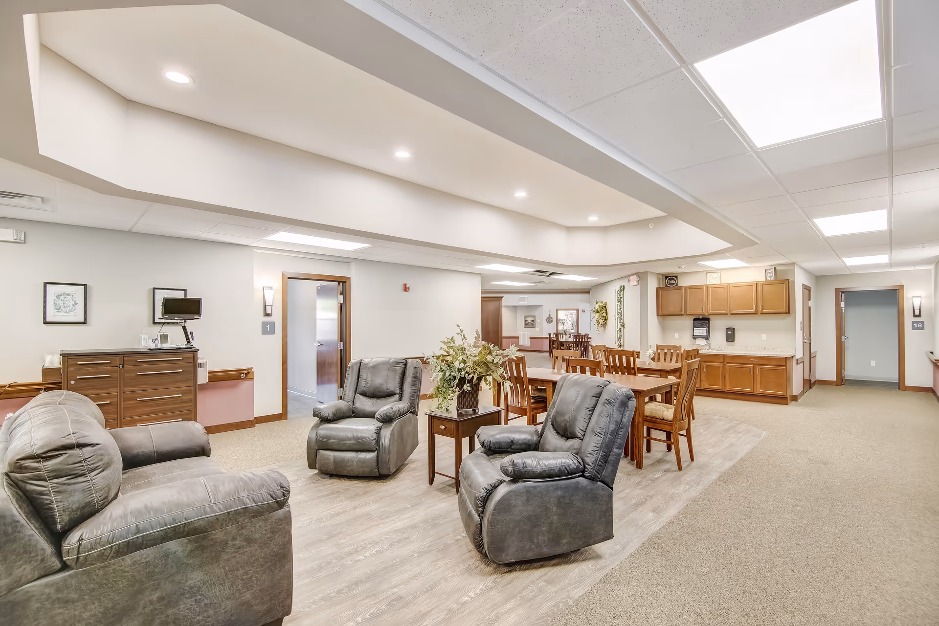 Spacious interior lounge and dining area with recliner chairs, wooden dining table, and a kitchenette in a senior living facility.