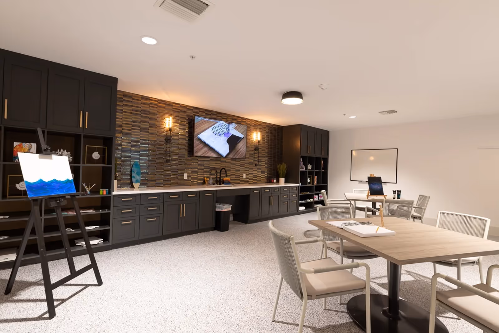 Well-lit interior activity room with tables and chairs, dark cabinetry and a tiled accent wall with a mounted TV, plus an easel with a painting.